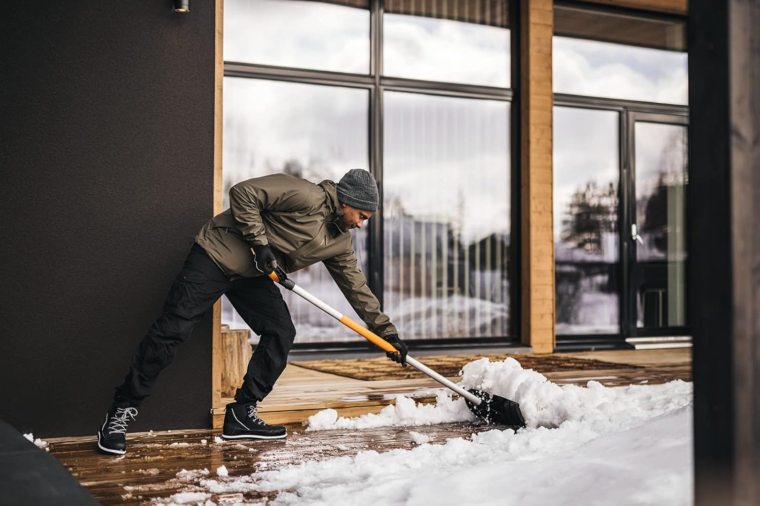 Uomo spalando neve su un patio in legno davanti a una casa moderna, indossando giacca invernale, pantaloni neri, guanti e berretto grigio. Scena invernale con attrezzo da neve, sfondo con finestre e paesaggio innevato.